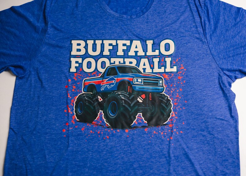 K&ouml;nnte beinhalten: Blaues T-Shirt mit dem Schriftzug "BUFFALO FOOTBALL" in wei&szlig;en Blockbuchstaben &uuml;ber einer Monster-Truck-Grafik. Der Truck ist blau mit roten und wei&szlig;en Akzenten und gro&szlig;en schwarzen Reifen. Rote, spritzerartige Akzente umgeben den Truck.