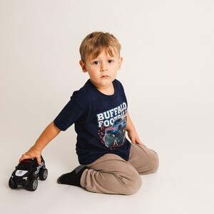 K&ouml;nnte beinhalten: Ein Kind tr&auml;gt ein marineblaues T-Shirt mit "BUFFALO FOOTBALL" und einem Monster-Truck-Motiv. Das Kind h&auml;lt ein schwarz-wei&szlig;es Polizei-Spielzeugauto. Das Kind tr&auml;gt eine beige Hose und schwarze Socken.
