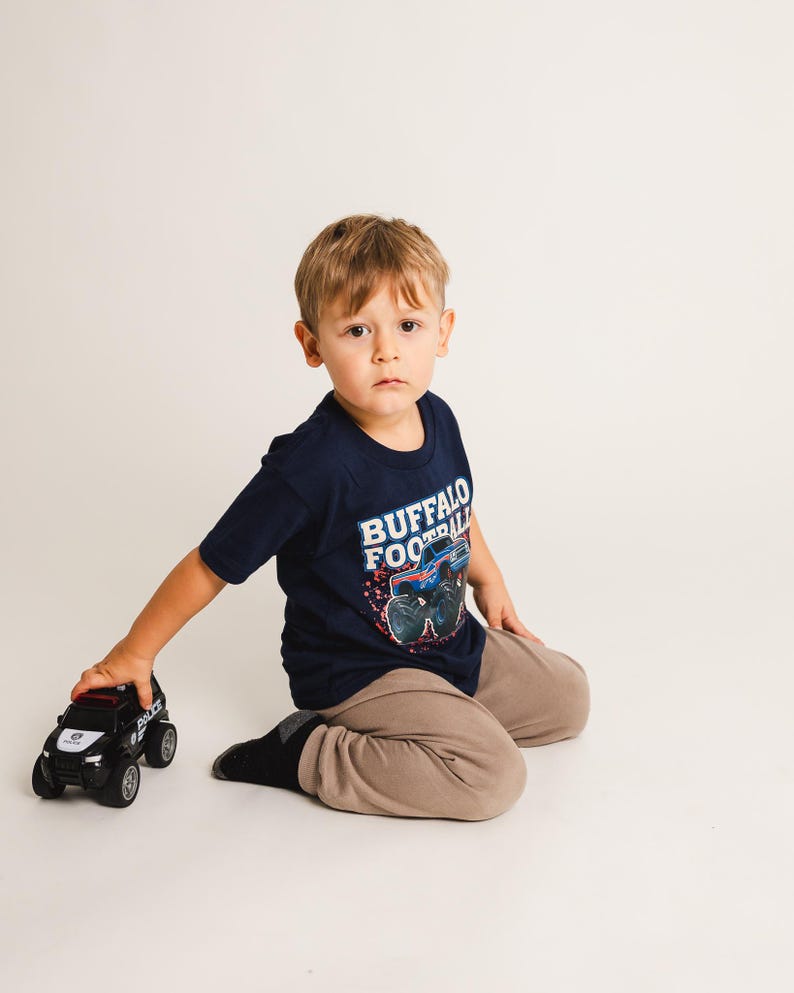 K&ouml;nnte beinhalten: Ein Kind tr&auml;gt ein marineblaues T-Shirt mit "BUFFALO FOOTBALL" und einem Monster-Truck-Motiv. Das Kind h&auml;lt ein schwarz-wei&szlig;es Polizei-Spielzeugauto. Das Kind tr&auml;gt eine beige Hose und schwarze Socken.