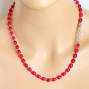 May include: A necklace featuring oval-shaped, deep red beads strung together with small silver-colored spacers. A section of the necklace is made of round, silver-colored beads. The necklace is displayed on a mannequin.