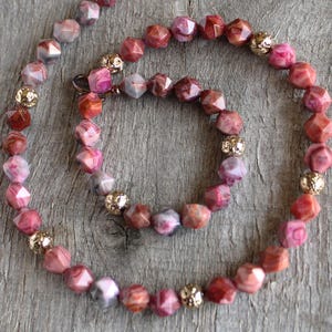 May include: A necklace with faceted, reddish-pink gemstone beads and gold-coloured, textured beads. The necklace is displayed on a wooden surface. The jewellery item features a mix of colours and textures. The necklace is a jewellery item.