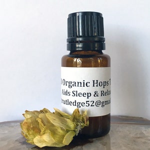 Puede incluir: Una pequeña botella de vidrio marrón con una tapa negra, etiquetada "Organic Hops Tincture Aids Sleep & Relax rutledge52@gmail.com". Un cono de lúpulo seco está al lado de la botella.