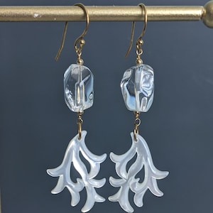 May include: A pair of drop earrings featuring gold-toned wire hooks, clear faceted rectangular beads, and white iridescent shell components carved into organic, branching, coral-like shapes that hang from the bottom of each clear bead.