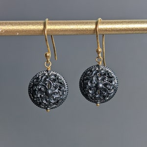 May include: A pair of black and gold filigree earrings with a floral design. The earrings are hanging from a gold bar.