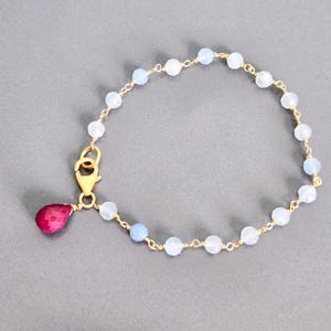 May include: A delicate gold-toned bracelet featuring alternating light blue and white beads. A teardrop-shaped, deep pink gemstone hangs from the clasp. The bracelet is designed with a wire-wrapped style.
