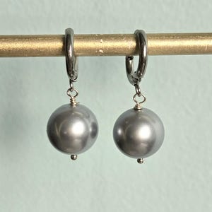 May include: A pair of silver-tone hoop earrings with large, gray, spherical pearl drops. The earrings hang from a gold-colored rod against a light blue background. The pearls have a glossy finish.