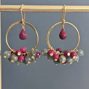 May include: Gold hoop earrings with a teardrop-shaped ruby gemstone dangling from each hoop. The hoops are adorned with a cluster of smaller ruby and labradorite gemstones.