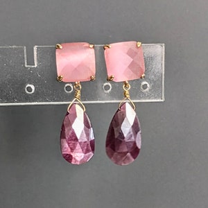 May include: A pair of pink and purple gemstone earrings. The earrings feature a square pink gemstone with a teardrop-shaped purple gemstone dangling below.