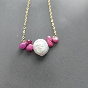May include: A gold chain necklace featuring a large, white, irregularly shaped pearl, accented with vibrant pink ruby gemstones. The necklace has a delicate design, with the gemstones clustered around the pearl.