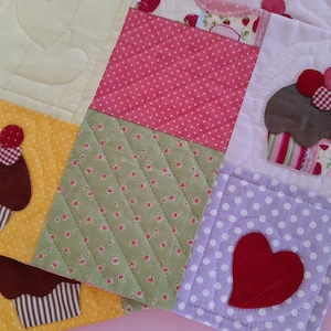 Peut inclure: Une courtepointe patchwork sur le thème des cupcakes. La courtepointe est faite de différents tissus, dont des pois roses, un imprimé floral vert et du blanc à pois violets. La courtepointe présente des cupcakes appliqués avec des cerises rouges et blanches.