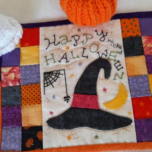 Può includere: Un runner trapuntato a tema Halloween con un bordo patchwork. Il centro presenta un cappello da strega, una ragnatela e le parole "Happy Halloween". Il bordo è un mix di quadrati arancioni, viola e fantasia.