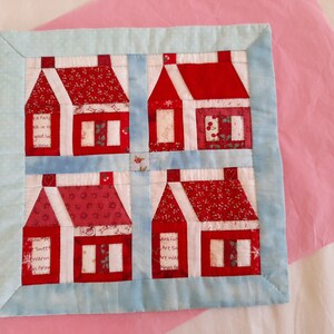 May include: A blue and white patchwork quilt with a design of four red houses. Each house has a white roof and windows, and the houses are arranged in a square pattern. The quilt is made with a variety of fabrics, including red, white, and blue.
