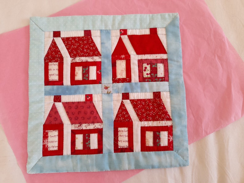 May include: A blue and white patchwork quilt with a design of four red houses. Each house has a white roof and windows, and the houses are arranged in a square pattern. The quilt is made with a variety of fabrics, including red, white, and blue.