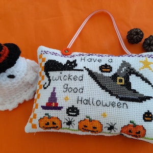 May include: A cross-stitch cushion with a Halloween theme, featuring the text "Have a wicked good Halloween." The design includes a black cat, witch's hat, pumpkins, and spiders. A small ghost and witch hat are to the left.