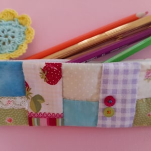 Op de afbeelding: Een patchwork etui met een rits. Het etui is gemaakt van verschillende stoffen, waaronder een lichtblauwe stof, een witte stof met aardbeien, een lichtgroene stof met stippen en een paars-wit geblokte stof. Het etui heeft twee knopen aan de voorkant.