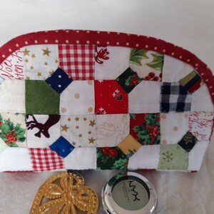 Può includere: Una pochette in tessuto patchwork rosso e bianco con la parte superiore curva. La pochette è realizzata con vari tessuti a tema natalizio, tra cui gingham rosso e bianco, agrifoglio verde e una stampa di renna. La pochette è rifinita con un tessuto rosso con pois bianchi.