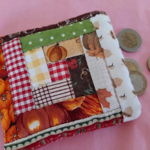 Puede incluir: Una pequeña cartera de monedas acolchada con un diseño de patchwork con colores y patrones otoñales, incluyendo calabazas, cuadros y hojas. La cartera está hecha de tela y tiene un fondo blanco con detalles marrones.
