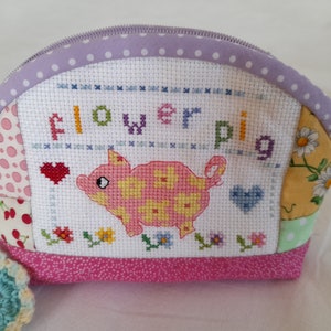May include: A pink and yellow pig with flower accents is embroidered on a white fabric pouch. The pouch is made of patchwork fabric with a pink, purple, and yellow floral design. The pouch has a zipper closure and is perfect for storing small items.