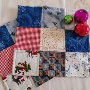 Könnte beinhalten: Zwei handgefertigte gequiltete Topflappen mit Weihnachtsmotiv. Die Topflappen bestehen aus Patchwork-Stoff mit rot-weißem Gingham, blau-weißen Schneeflocken und einem weißen Hintergrund mit einem Weihnachtsbaum- und Hausdesign. Die Topflappen sind mit roten und grünen Ornamenten verziert.