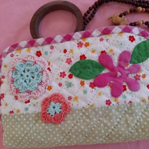 Può includere: Una pochette trapuntata floreale con manico in legno. La pochette presenta uno sfondo bianco con piccoli fiori, fiori all'uncinetto e un grande fiore rosa. La sezione inferiore è verde chiaro con pois bianchi.