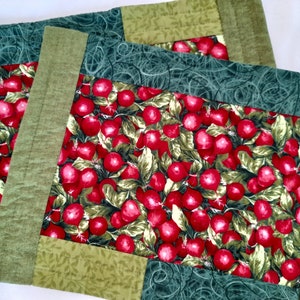 Puede incluir: Dos manteles individuales acolchados verdes y rojos con estampado de cerezas. Los manteles individuales tienen un borde verde y un patrón de hojas verdes y blancas.
