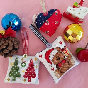 Può includere: Una collezione di decorazioni natalizie, tra cui una sfera blu sfaccettata, una pigna con un fiocco rosso e una slitta rossa e bianca con una figura di Babbo Natale. Altri ornamenti includono un cuore, una palla da discoteca dorata e cuscini ricamati.