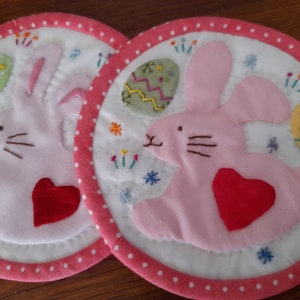 Pode incluir: Dois  coasters de tecido branco com coelhos cor-de-rosa segurando corações vermelhos. Cada coelho tem um ovo bordado com um desenho amarelo, verde e branco.