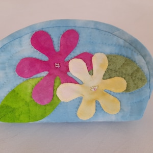 Può includere: Una pochette in tessuto trapuntato di colore azzurro chiaro con chiusura a zip. La pochette è decorata con due fiori in tessuto, uno rosa e uno giallo, con foglie in tessuto verde.