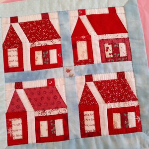 May include: A red and white patchwork quilt with a design of four houses. The houses are made of different fabrics with various patterns and have white trim. The quilt is bordered with a light blue fabric.