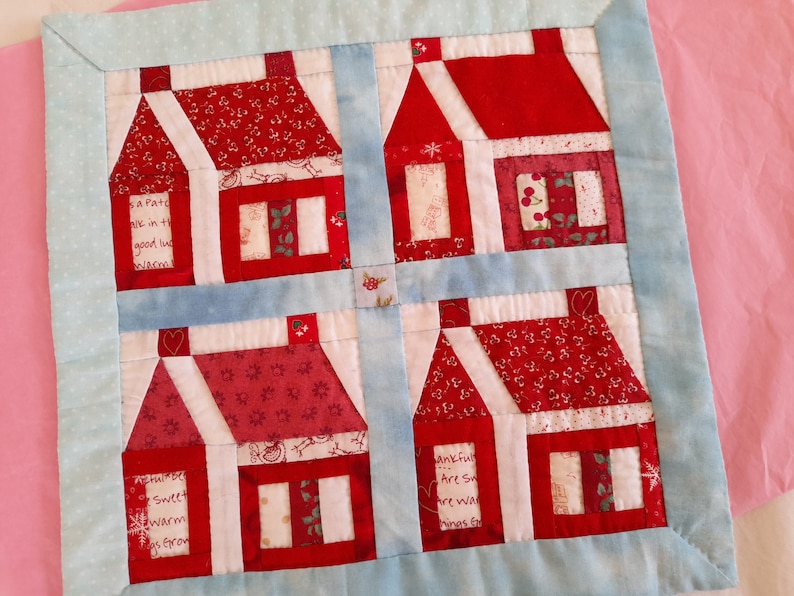 May include: A red and white patchwork quilt with a design of four houses. The houses are made of different fabrics with various patterns and have white trim. The quilt is bordered with a light blue fabric.