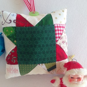 May include: A handmade Christmas ornament in the shape of a pillow with a patchwork design featuring red, green, white, and gold fabrics. The pillow has a green button and a loop for hanging.
