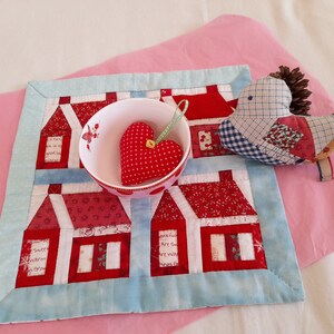 May include: A blue and white quilted fabric with red houses and a red heart in a white bowl. A patchwork bird with a brown pinecone on its head is on the fabric.