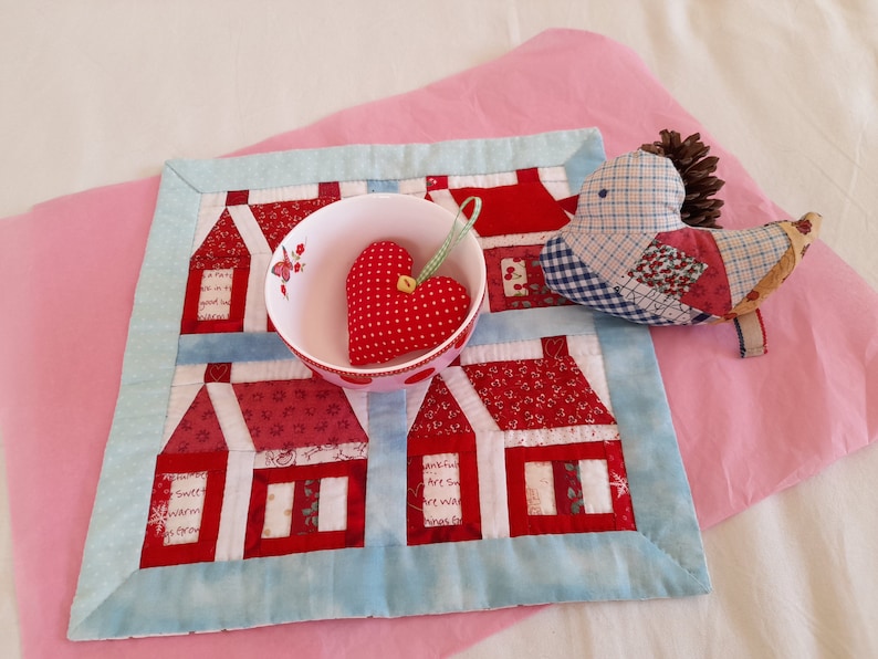 May include: A blue and white quilted fabric with red houses and a red heart in a white bowl. A patchwork bird with a brown pinecone on its head is on the fabric.