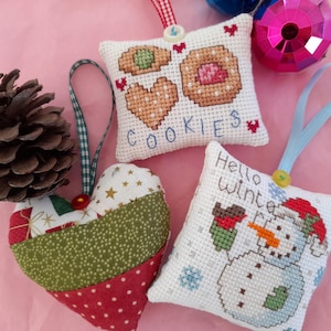Op de afbeelding: Een verzameling handgemaakte kerstversieringen. De afbeelding toont een hartvormige ornament, een dennenappel en twee vierkante ornamenten met kruissteekontwerpen. Eén ornament zegt "Cookies", de andere "Hello Winter".