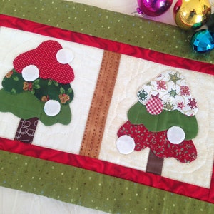 May include: A white quilted table runner with a red and green border. The runner features two appliqued Christmas trees with white ornaments. The trees are made from different patterned fabrics.