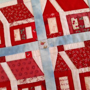 May include: A quilt block with a patchwork design featuring four red and white houses with different patterns. The houses are surrounded by a blue border.