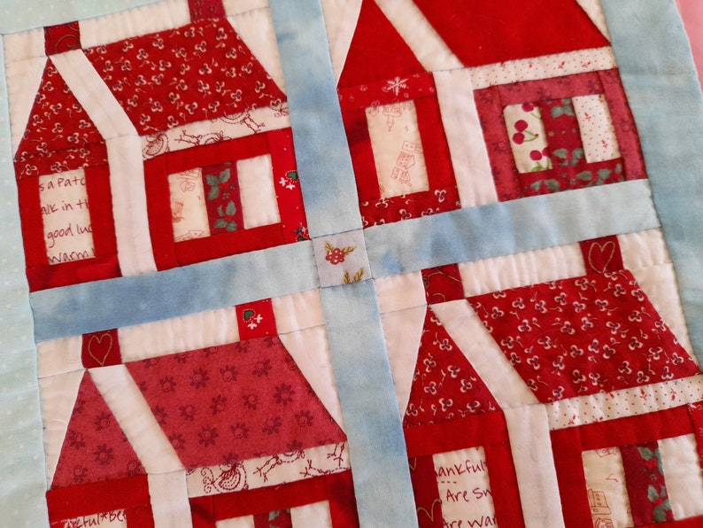 May include: A quilt block with a patchwork design featuring four red and white houses with different patterns. The houses are surrounded by a blue border.