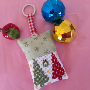 Può includere: Un portachiavi natalizio fatto a mano con un cuscino ricamato a punto croce con alberi di Natale verdi e rossi e una ghirlanda. Include un ornamento a forma di mela rossa e due ornamenti sfaccettati.