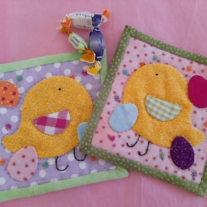 Può includere: Due sottobicchieri quadrati in tessuto trapuntato con applicazioni di uccelli gialli, ciascuno con un uovo a fantasia diverso. Uno ha uno sfondo lavanda con pois bianchi, l'altro ha uno sfondo rosa con piccoli motivi floreali. Caramelle sullo sfondo.