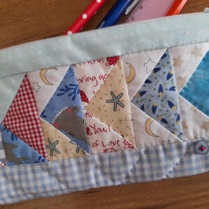 Może przedstawiać: Niebieska i biała kosmetyczka w patchworku z zamkiem błyskawicznym. Kosmetyczka jest wykonana z różnych tkanin o różnych wzorach, w tym gwiazd, księżyców i kratki. Kosmetyczka ma zapięcie na guzik i małą kieszeń z przodu.