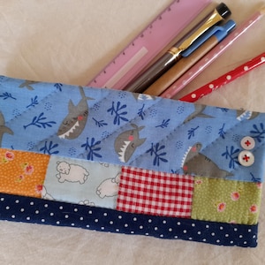 Könnte beinhalten: Ein gestepptes Federmäppchen mit blauem Hai-Muster, Patchwork-Design und einem marineblauen Polka-Dot-Rand. Das Etui enthält ein rosa Lineal, Stifte und einen rot gepunkteten Bleistift. Zwei weiße Knöpfe mit roten Kreuzen befinden sich an der Seite.