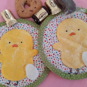 Può includere: Due sottobicchieri rotondi in tessuto verde e bianco con un design di pulcino giallo. Ogni sottobicchiere ha una forma di uovo bianco e un motivo a coriandoli colorati.