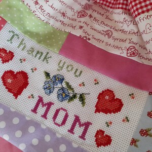 Könnte beinhalten: Ein weißer Stoff mit einem Kreuzstich-Design, auf dem "Danke MAMA" mit roten Herzen und blauen Blumen steht. Der Stoff ist von einem Patchwork aus verschiedenen Stoffen mit Punkten, Karos und Blumenmustern umgeben.