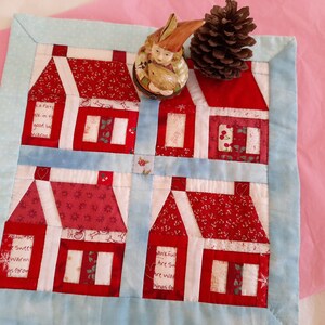 May include: A blue and white quilted wall hanging with a red and white patchwork design of four houses. The houses have white windows and red roofs. The houses are surrounded by a blue border. The wall hanging is on a pink background.