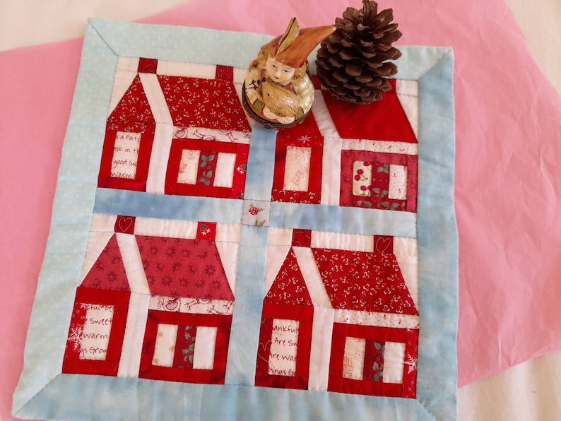 May include: A blue and white quilted wall hanging with a red and white patchwork design of four houses. The houses have white windows and red roofs. The houses are surrounded by a blue border. The wall hanging is on a pink background.