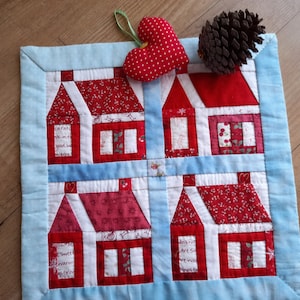 May include: A blue and red patchwork quilt with a design of four houses. The houses are made of different fabrics and have white trim. The quilt is bordered with a light blue fabric. A red heart with white polka dots is in the upper right corner of the quilt.