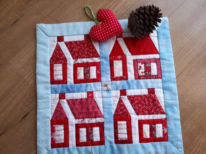 May include: A blue and red patchwork quilt with a design of four houses. The houses are made of different fabrics and have white trim. The quilt is bordered with a light blue fabric. A red heart with white polka dots is in the upper right corner of the quilt.