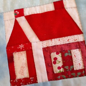 May include: A red and white patchwork quilt block featuring a house design. The house has a red roof, white walls, and a red door. The quilt block is made up of various fabrics with different patterns, including polka dots, snowflakes, and cherries.