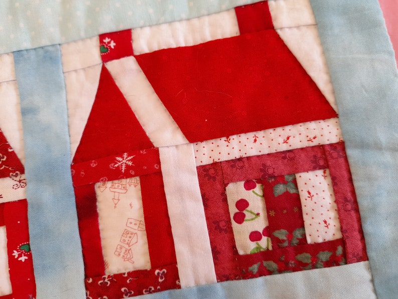 May include: A red and white patchwork quilt block featuring a house design. The house has a red roof, white walls, and a red door. The quilt block is made up of various fabrics with different patterns, including polka dots, snowflakes, and cherries.