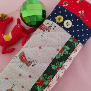 May include: A quilted eyeglass case with a patchwork design featuring red, blue, green, and white fabrics. The case has a reindeer pattern and a holly pattern. There are two buttons on the case.